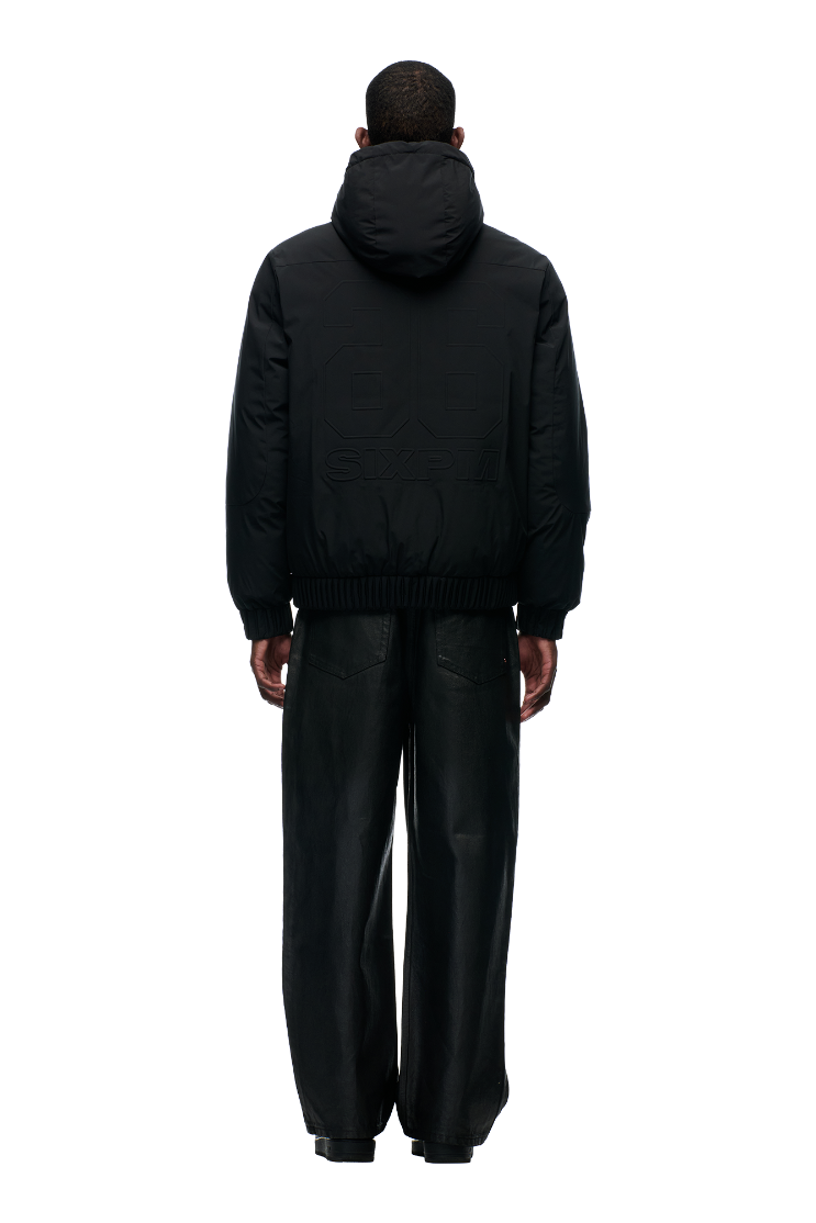 A person stands facing away, wearing the 6PM EMBOSSED JACKET BLACK with a hidden arm pocket and loose black pants, against a plain light gray background.