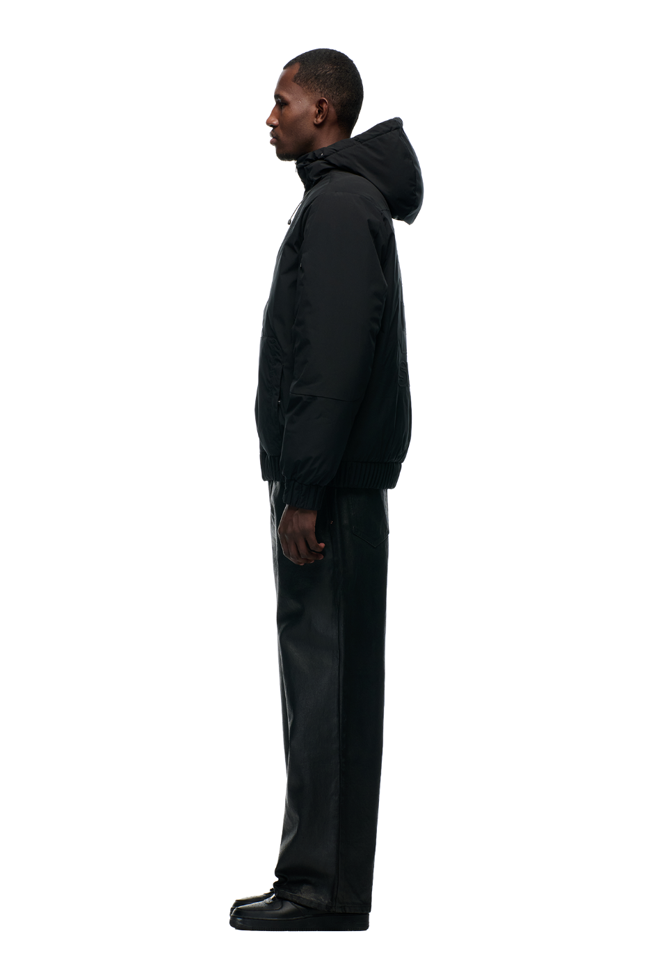 A man stands in profile against a plain background, wearing the 6PM EMBOSSED JACKET BLACK with a hidden arm pocket, paired with black pants and shoes.