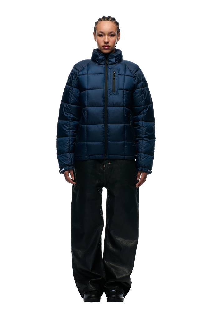 A person faces forward against a plain light background, wearing the 6PM LIGHT PUFFER JACKET RECTANGLES MIDNIGHT BLUE paired with black baggy pants.