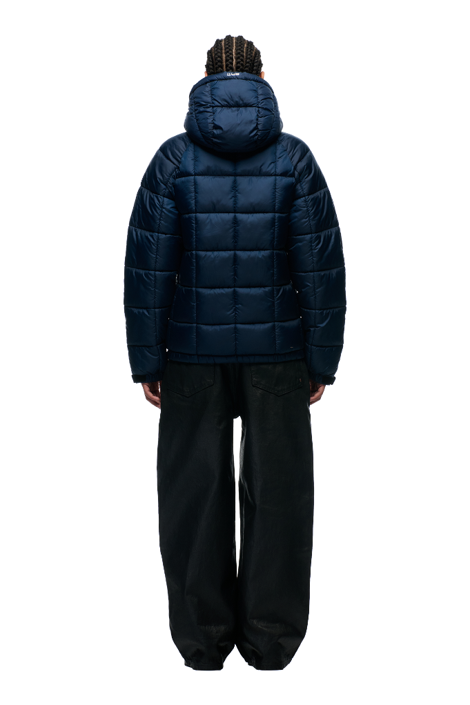 A person with braided hair is seen from behind, wearing the 6PM LIGHT PUFFER JACKET RECTANGLES MIDNIGHT BLUE and loose-fitting black pants, against a plain light background.