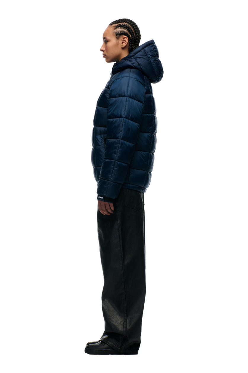 A person with braided hair stands in profile wearing the 6PM LIGHT PUFFER JACKET RECTANGLES MIDNIGHT BLUE, wide-leg black pants, and black shoes against a plain white background.
