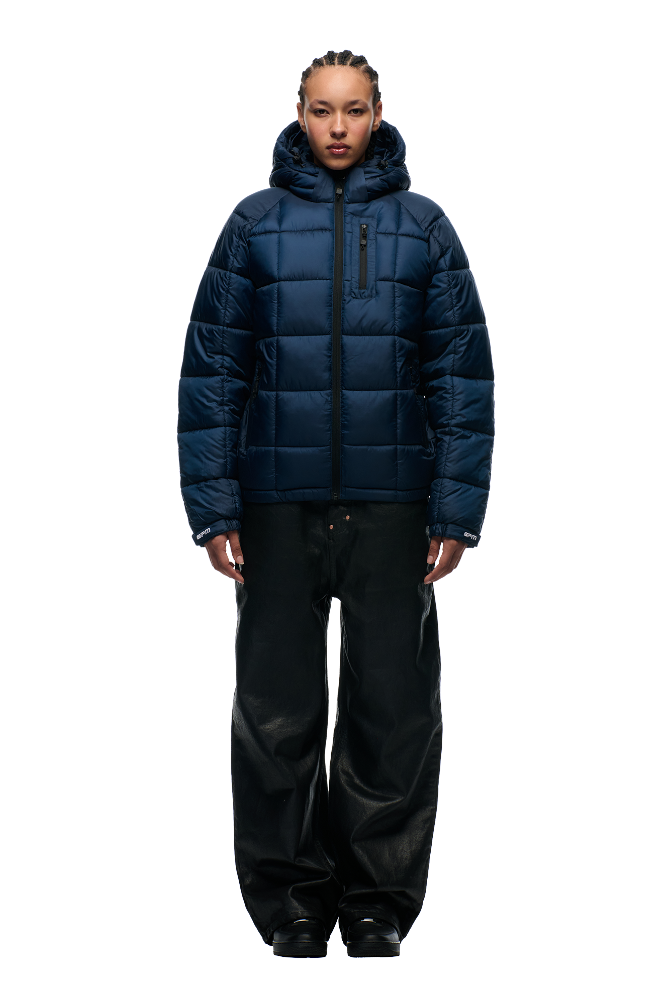 A person stands facing forward, wearing the 6PM LIGHT PUFFER JACKET RECTANGLES MIDNIGHT BLUE with an adjustable hood and black wide-leg pants against a plain, light-colored background.