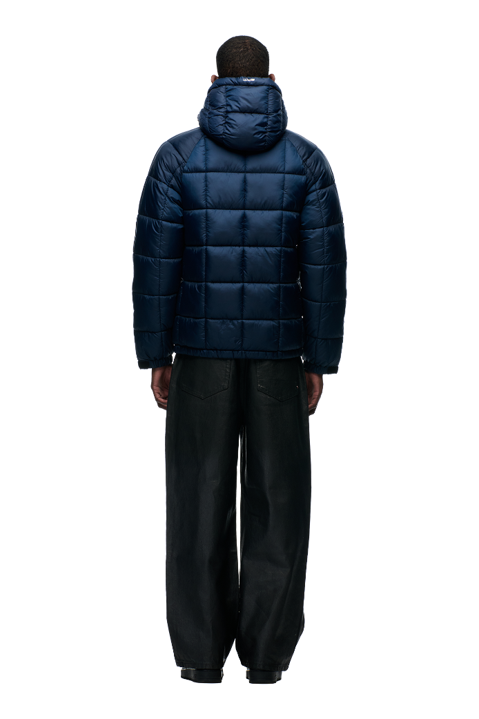 A person stands facing away against a white background, wearing the 6PM LIGHT PUFFER JACKET RECTANGLES in midnight blue with an adjustable hood and loose black pants.