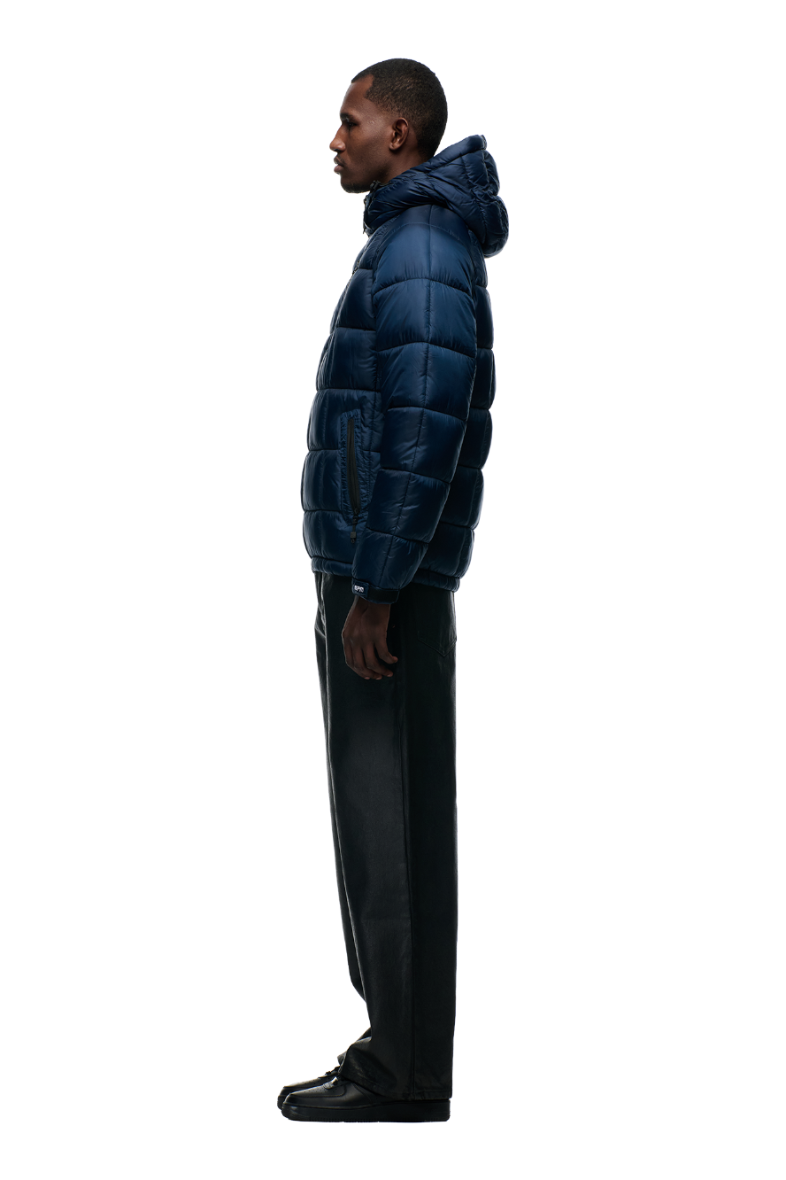 A man stands in profile against a plain background, wearing the 6PM LIGHT PUFFER JACKET RECTANGLES MIDNIGHT BLUE, paired with black pants and black shoes.