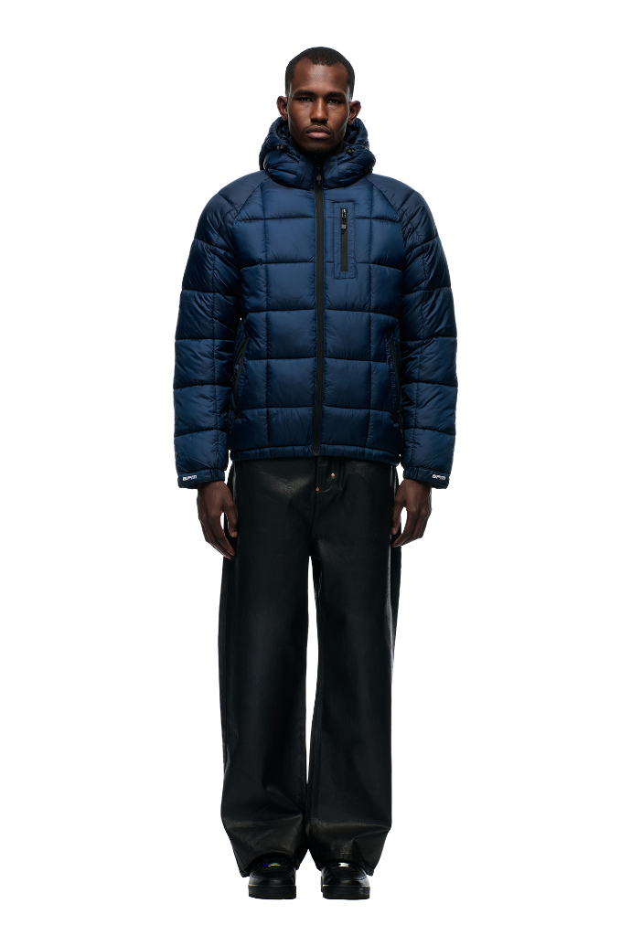 A man faces forward against a plain white background wearing the 6PM LIGHT PUFFER JACKET RECTANGLES MIDNIGHT BLUE, paired with black wide-leg pants and black shoes.
