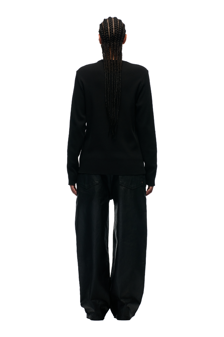 A person with long, braided hair stands facing away, wearing the 6PM 6PM+ RIBBED LS BLACK top and loose-fitting black pants against a plain white background.
