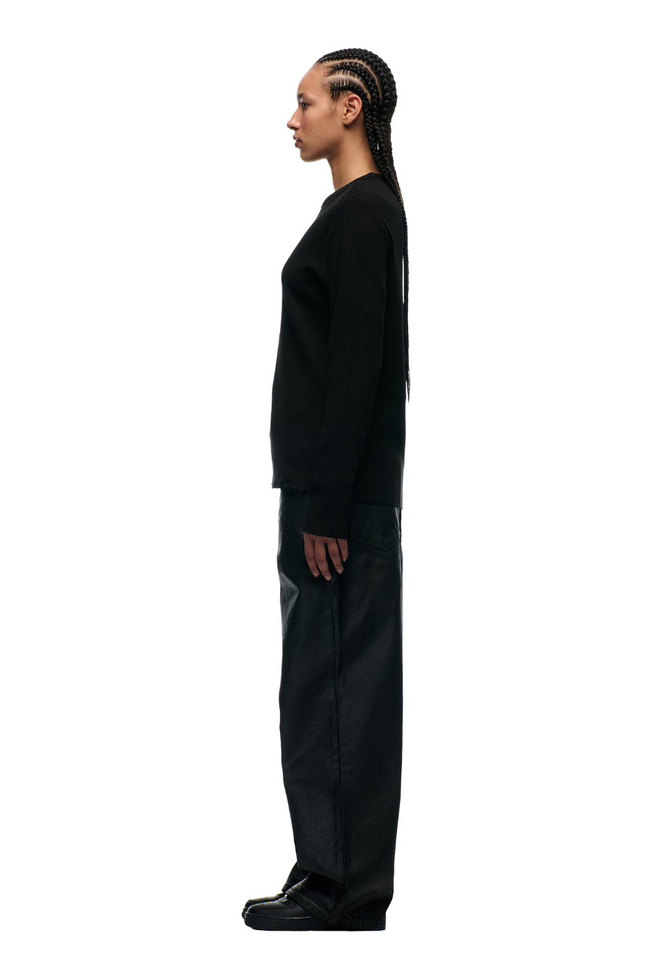 A person with long braided hair stands in profile facing left, wearing the 6PM RIBBED LS BLACK top from 6PM, loose black pants, and black shoes with a tonal flag label against a plain white background.