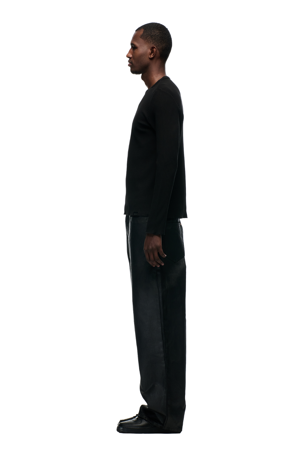 A man stands in profile against a white background, wearing the 6PM 6PM+ RIBBED LS BLACK top, dark loose-fitting pants, and black shoes, with a tonal flag label adding a subtle detail.
