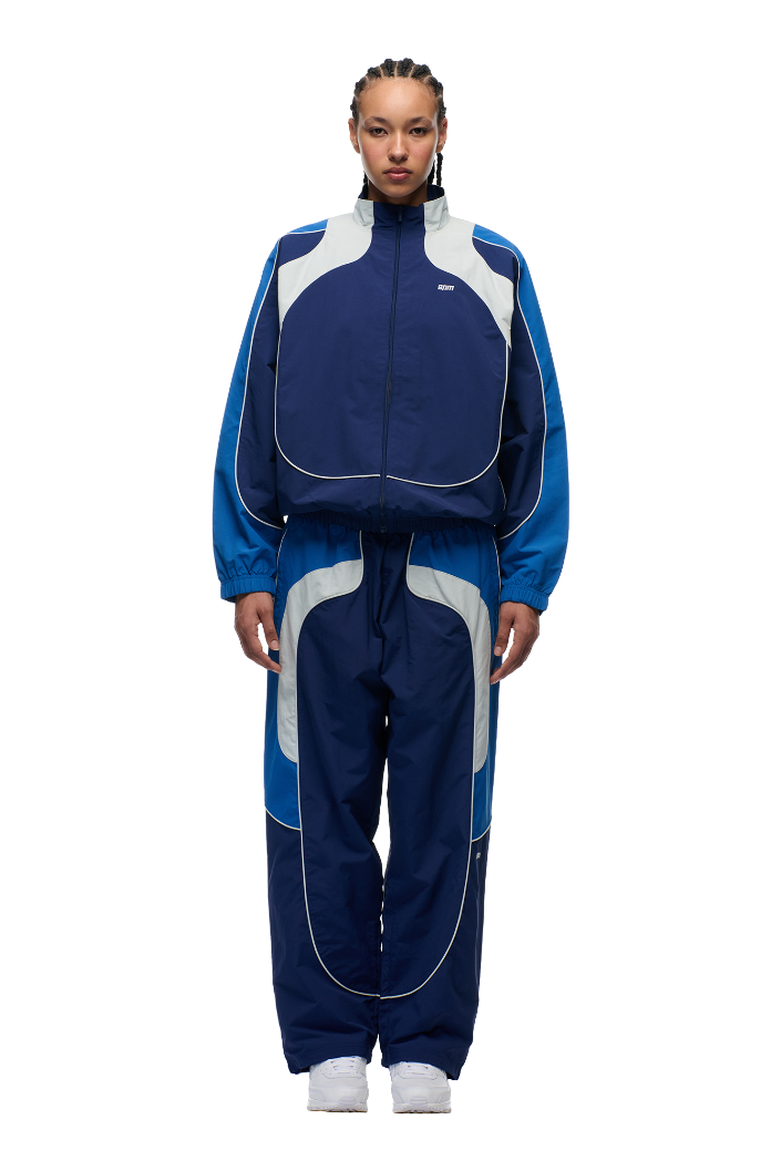 A person with braided hair models the 6PM EVERYDAY TRACK JACKET SHADOW BLUE—a loose-fitting, color-blocked navy, light blue, and gray nylon jacket—standing facing forward against a plain white background.