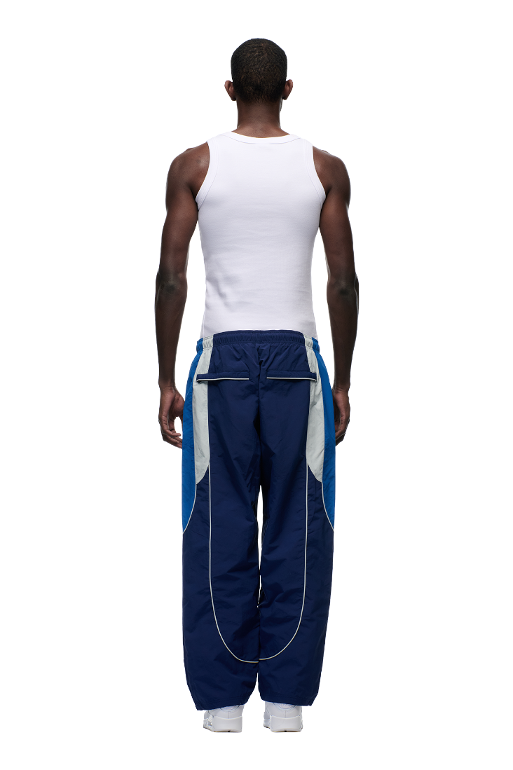 A man faces away from the camera, wearing 6PM’s EVERYDAY TRACK PANTS SHADOW BLUE with light blue and gray panels, zipped pockets, a white tank top, and white sneakers, set against a plain white background.