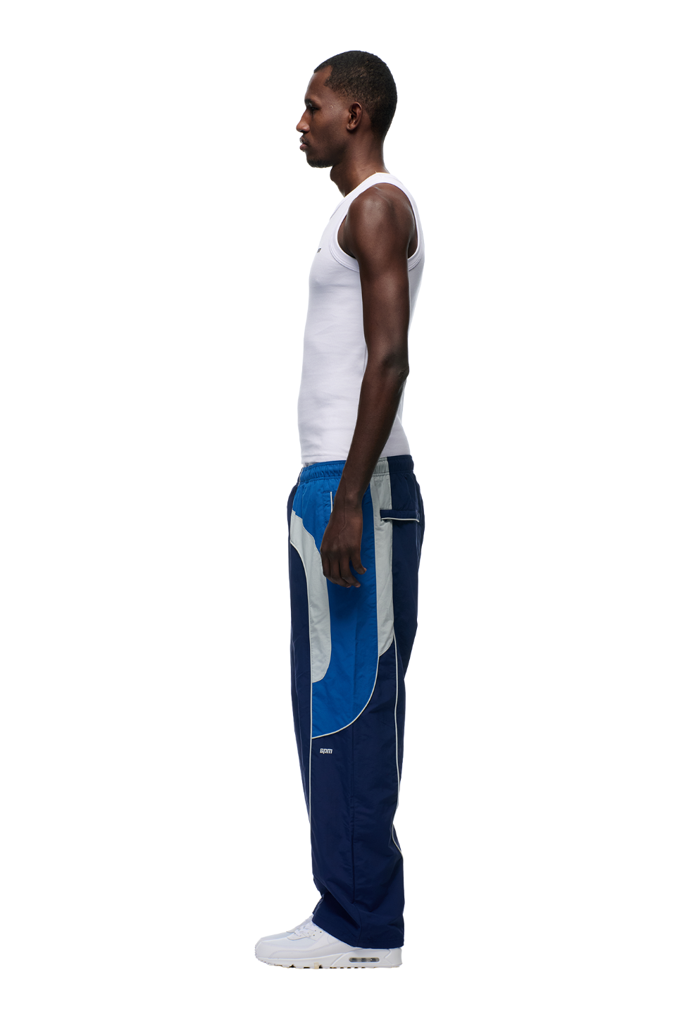 A man stands in profile facing left on a plain white background, wearing a white sleeveless tank top, 6PM EVERYDAY TRACK PANTS SHADOW BLUE with white and light blue panels plus zipped pockets, and white sneakers.