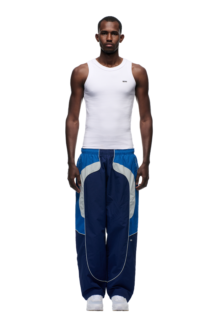 A man faces forward against a plain white background, wearing a fitted white tank top, white sneakers, and 6PM’s EVERYDAY TRACK PANTS SHADOW BLUE with white and gray curved accents and zipped pockets.