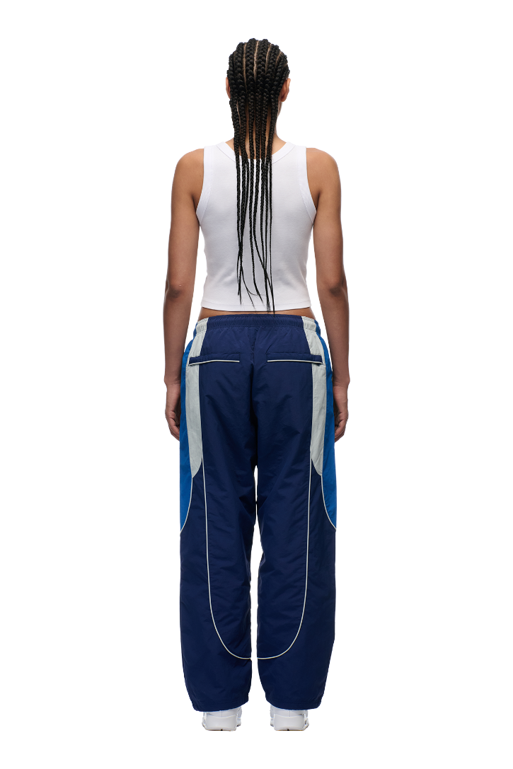A person with long braids, in a white tank top and 6PM EVERYDAY TRACK PANTS SHADOW BLUE featuring gray and blue accents and zipped pockets, stands facing away against a white background.