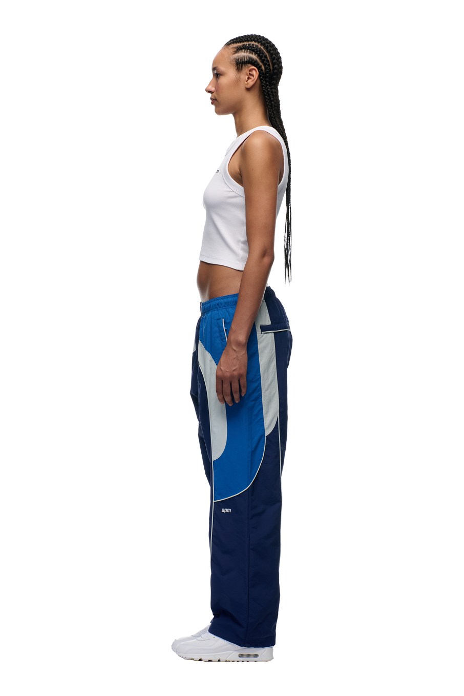 A woman with long braids stands in profile against a plain white background, wearing a white tank top, 6PM EVERYDAY TRACK PANTS SHADOW BLUE with zipped pockets, and white sneakers.