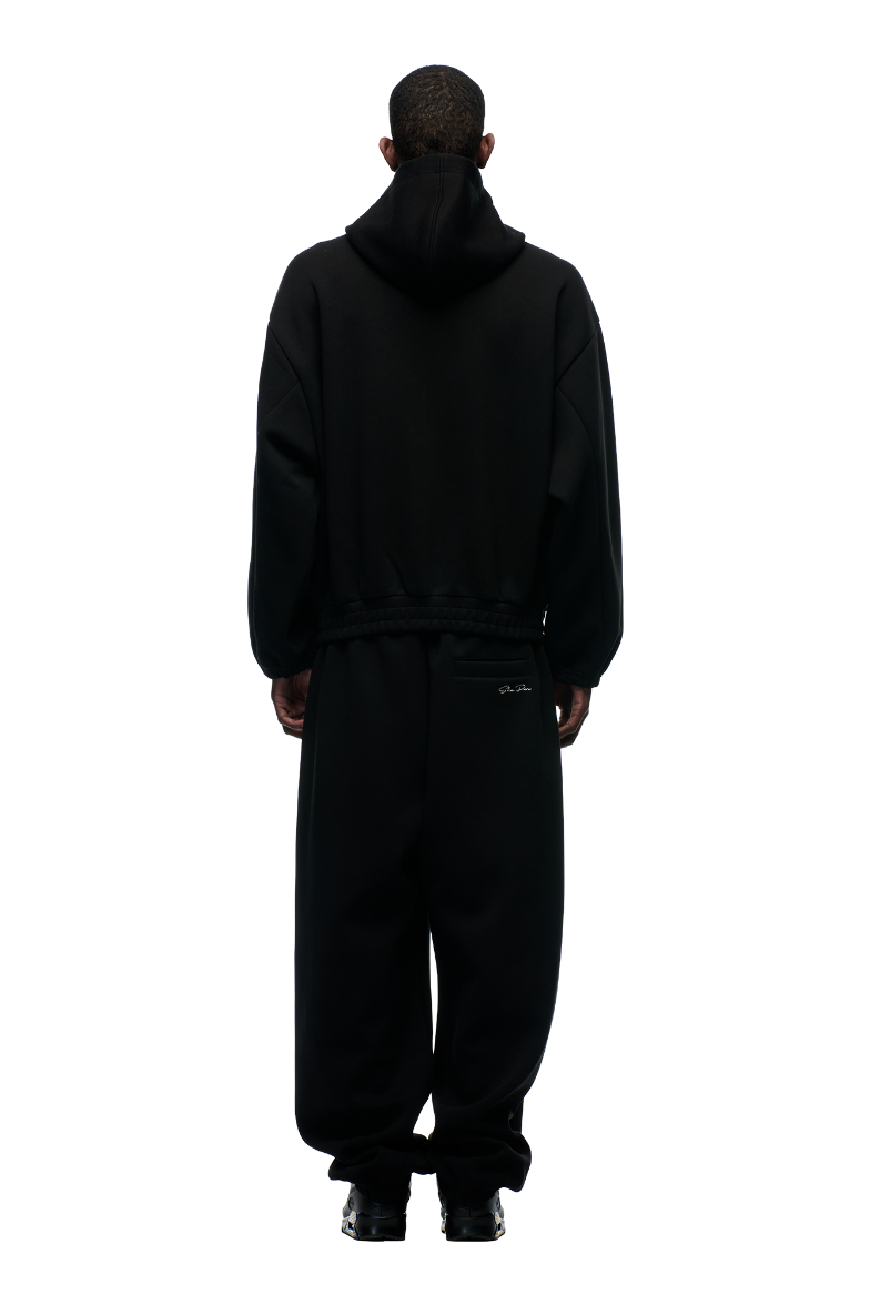 A person faces away from the camera, wearing the 6PM BOXY ZIP HOODIE BLACK with a metal zipper and matching black pants, set against a plain light background.