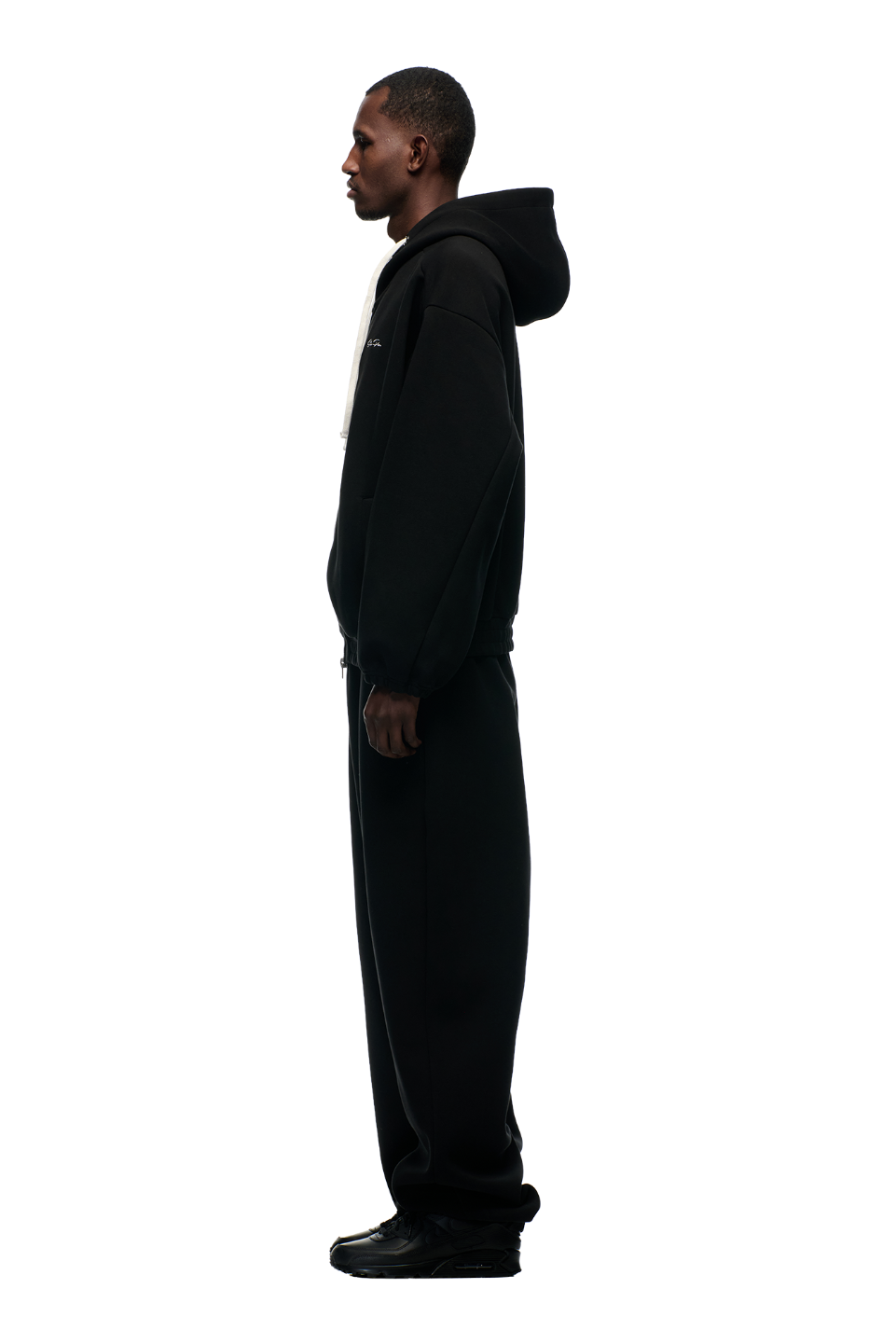 A man stands in profile facing left, wearing the 6PM BOXY ZIP HOODIE BLACK with a metal zipper, loose black sweatpants, and black shoes against a plain white background.