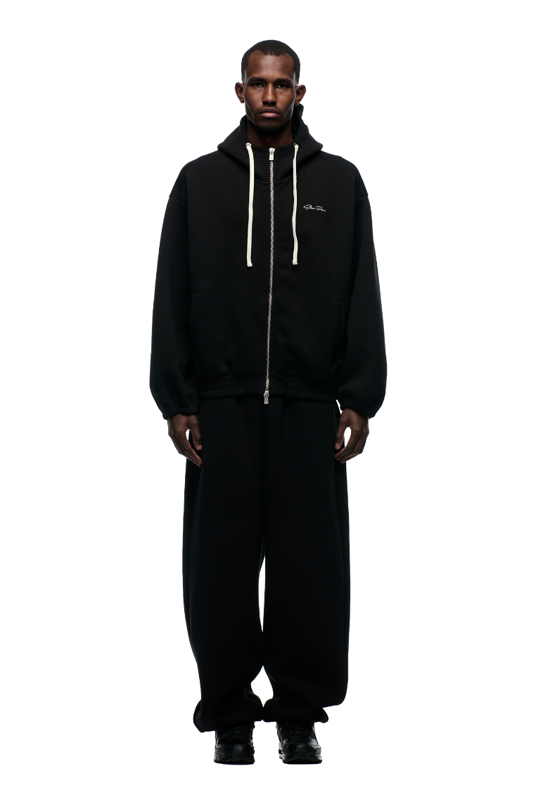 A man stands facing forward against a plain light background, wearing the 6PM BOXY ZIP HOODIE BLACK with white drawstrings, embroidered logo, metal zipper, and matching wide-leg black pants and black shoes.