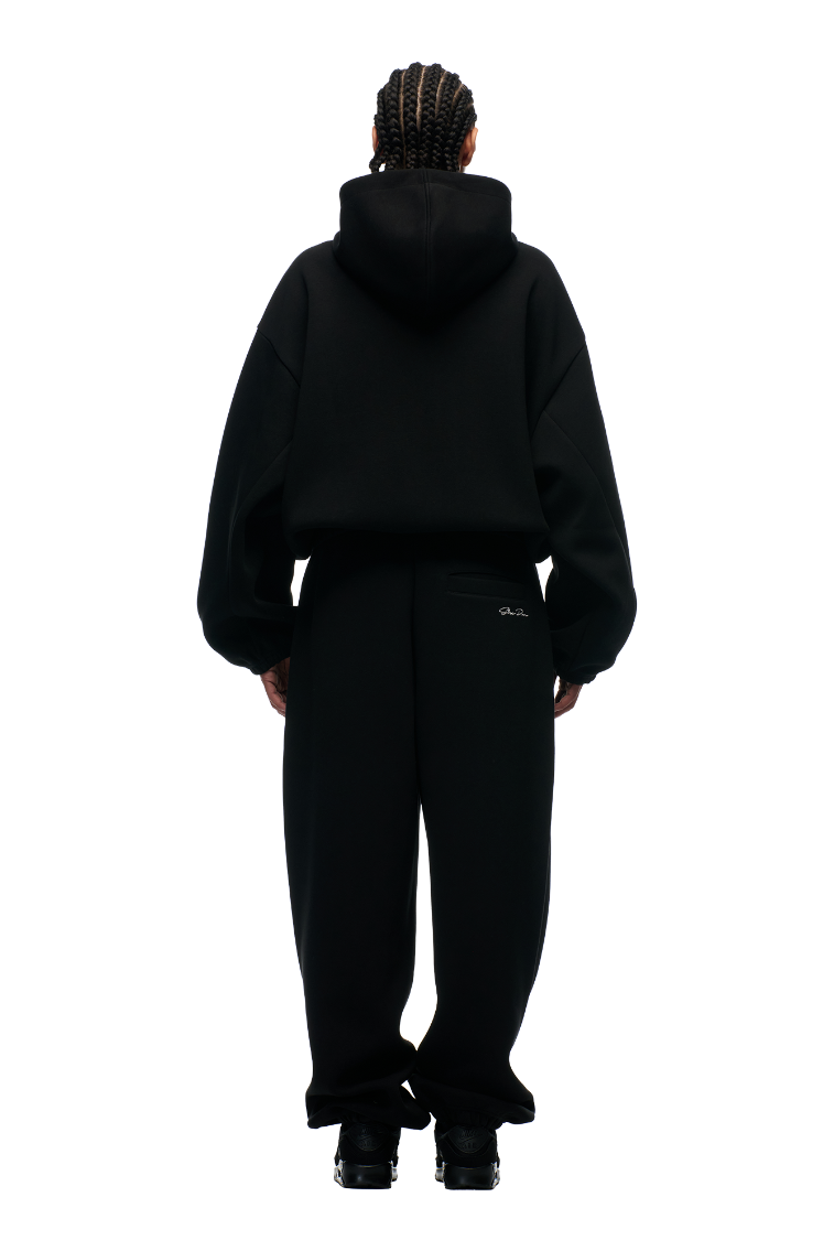 A person with braided hair faces away, wearing the 6PM BOXY ZIP HOODIE BLACK with a metal zipper, paired with black pants and shoes, against a plain white background.