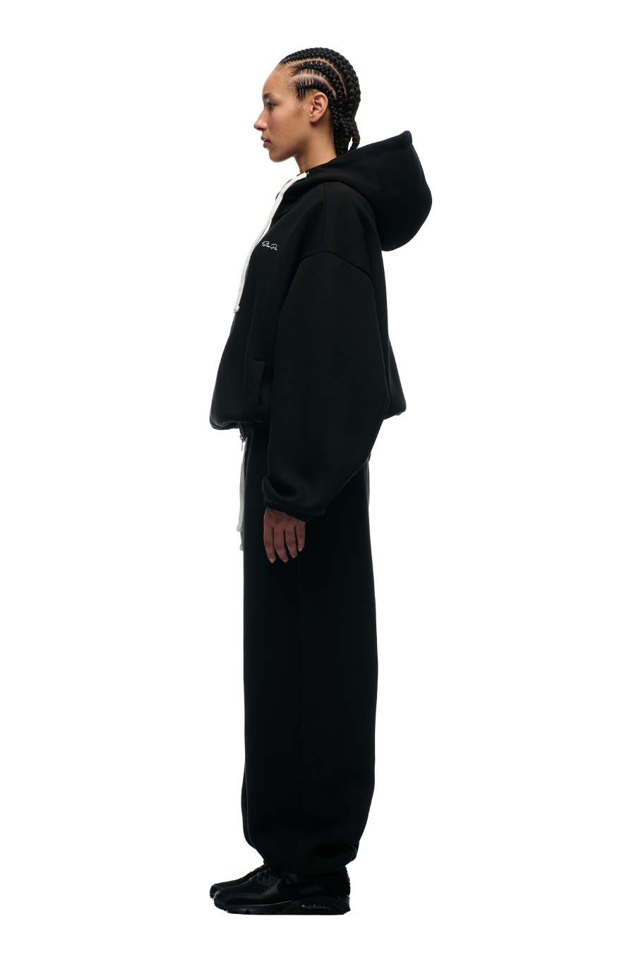 A person with braided hair stands in profile wearing the 6PM BOXY ZIP HOODIE BLACK, paired with loose black pants and black shoes, set against a plain light background.