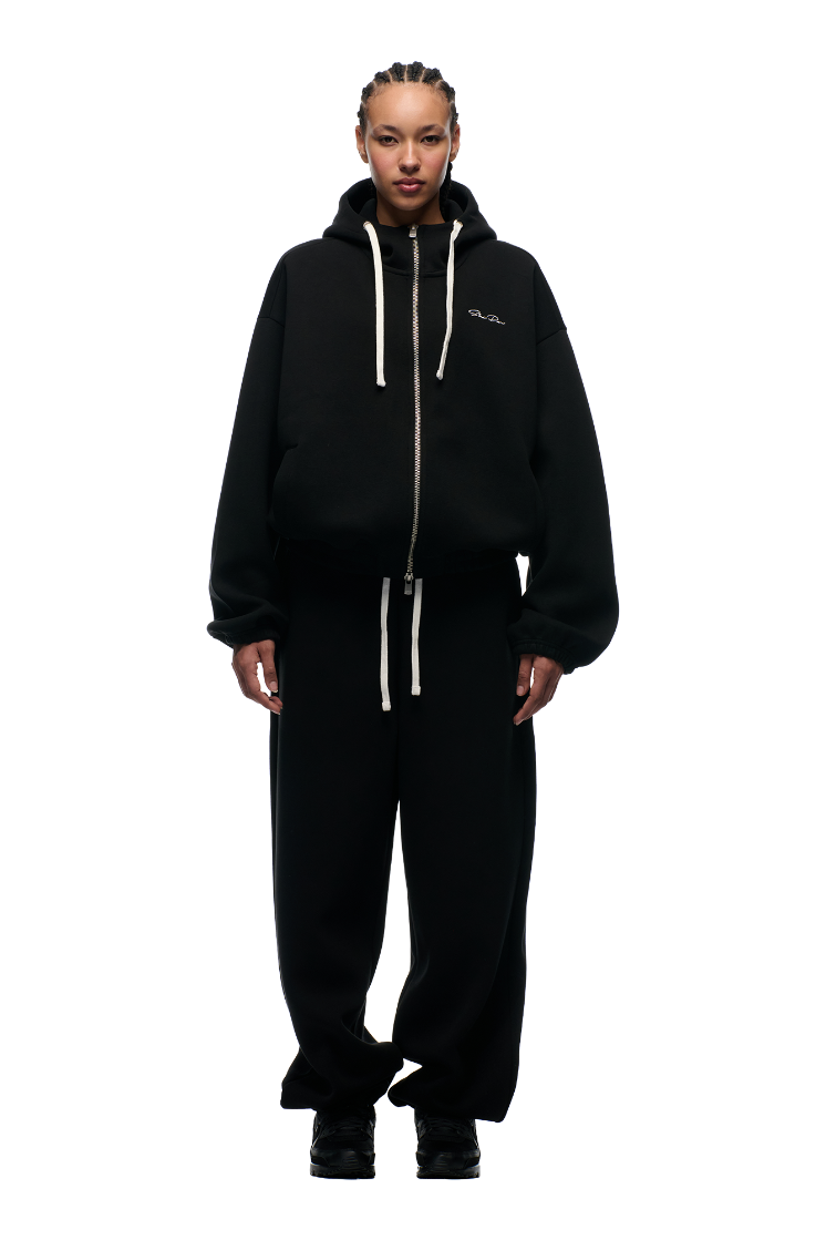 A person faces forward, wearing the 6PM BOXY ZIP HOODIE BLACK with white drawstrings and black baggy pants. They have braided hair, black shoes, and a neutral expression against a plain light background.