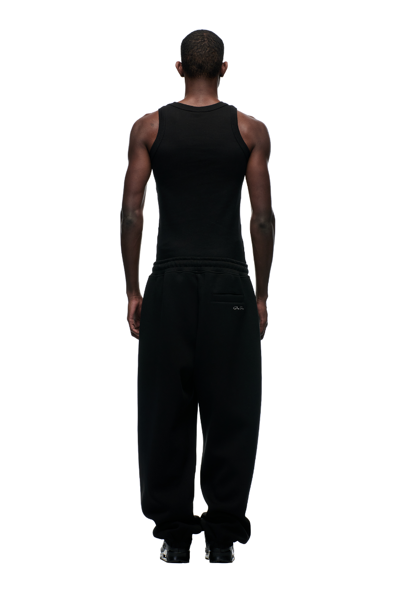 A person stands with their back to the camera, wearing 6PM's X-TRA WIDE CUFF JOGGER BLACK, a black tank top, and black shoes against a plain white background.