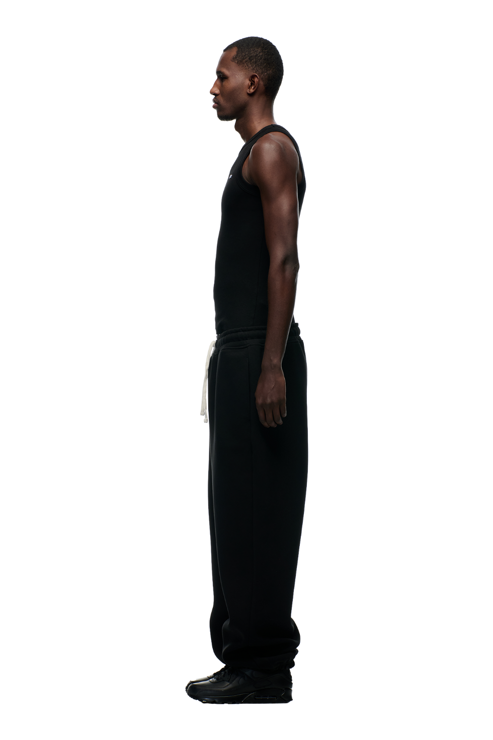 A man in a fitted black tank top, 6PM X-TRA WIDE CUFF JOGGER BLACK sweatpants with a white drawstring, and black sneakers stands in profile against a plain white background.