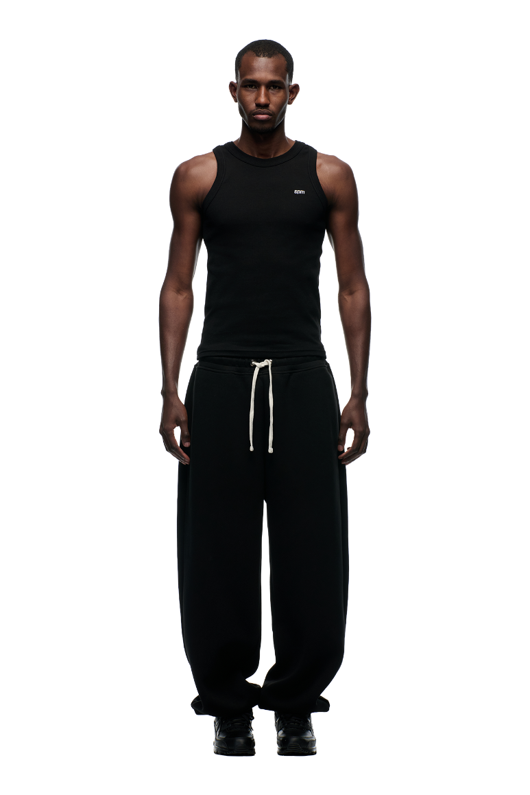A man stands facing forward in a fitted black tank top and 6PM X-TRA WIDE CUFF JOGGER BLACK pants with white laces, wide cuffs, and black shoes, set against a plain white background.