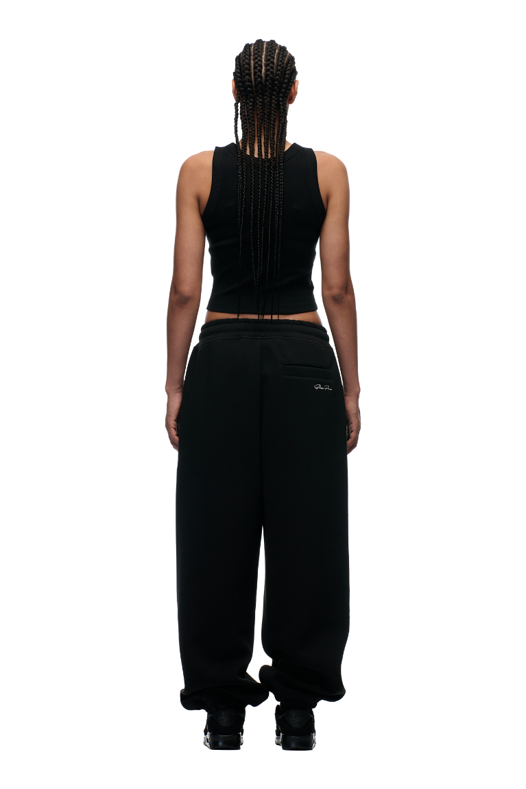 A person with long braids, dressed in a black sleeveless top and 6PM's X-TRA WIDE CUFF JOGGER BLACK pants, stands facing away against a plain white background.