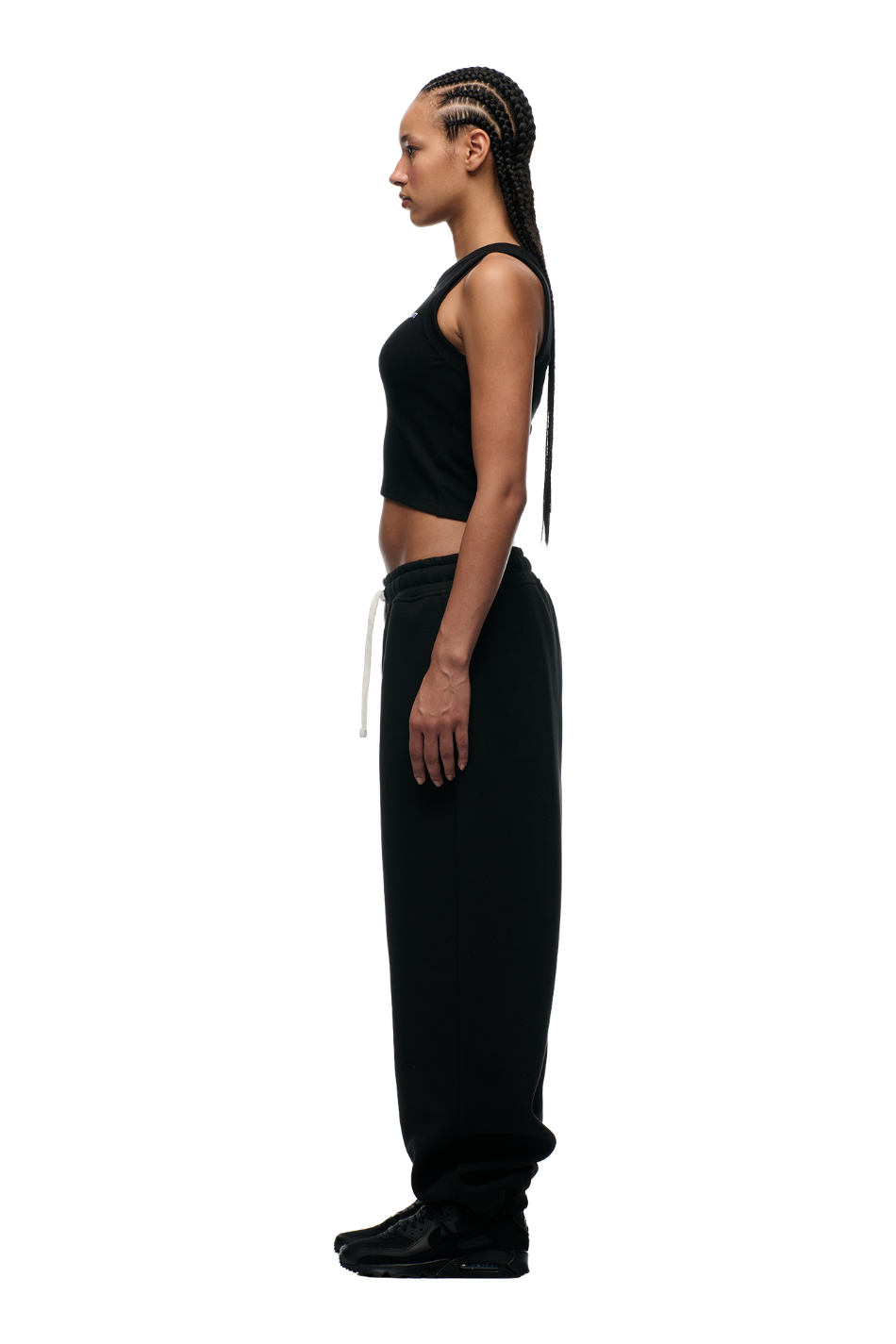 A woman with long braided hair stands in profile against a white background, wearing a black sleeveless top, 6PM X-TRA WIDE CUFF JOGGER BLACK sweatpants, and black sneakers.