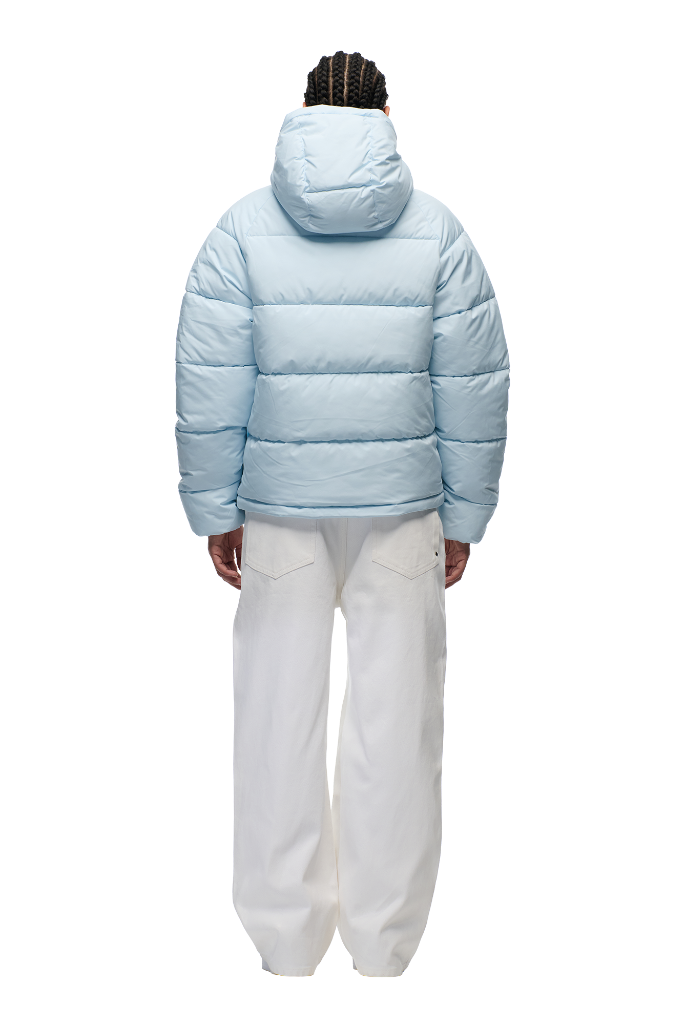 A person with braided hair, seen from behind, wears the 6PM PUFFER JACKET ICE BLUE with an adjustable hood and loose white pants, standing against a plain white background.