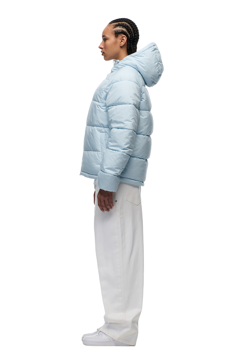 A person stands in profile against a white background, wearing the 6PM PUFFER JACKET ICE BLUE with an adjustable hood, paired with white pants and sneakers.