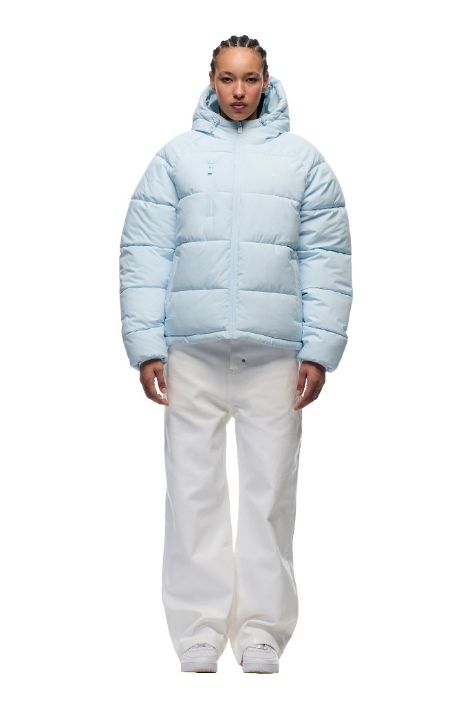 A person faces forward against a plain white background, wearing the 6PM PUFFER JACKET ICE BLUE with an adjustable hood, paired with white wide-leg pants and white sneakers.