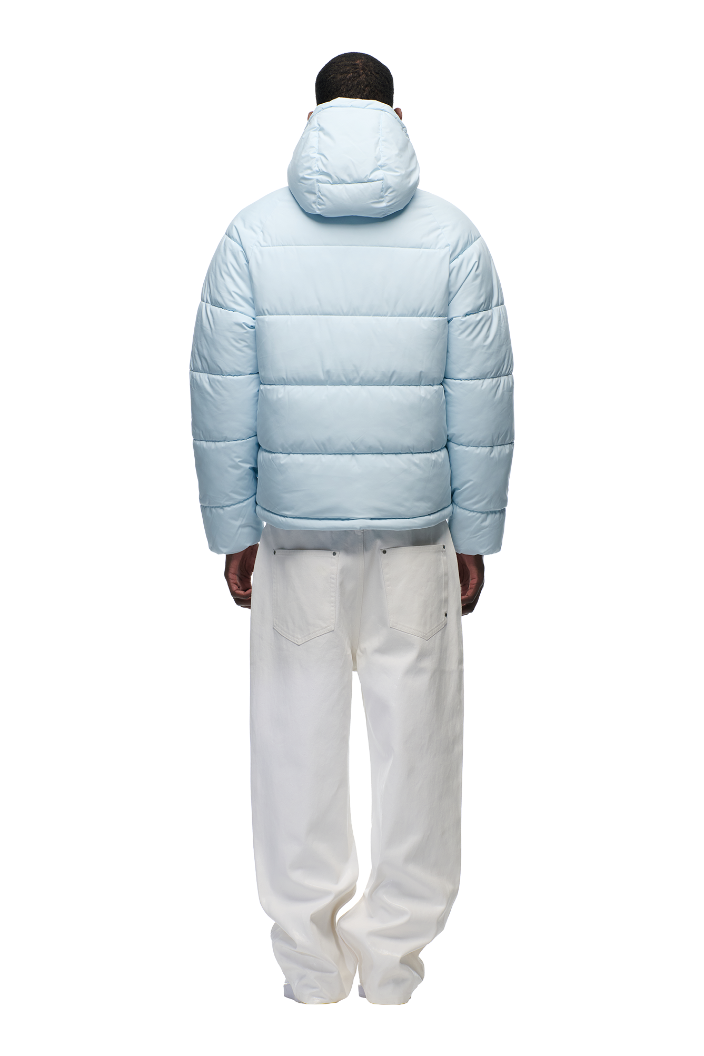 A person faces away, wearing the 6PM PUFFER JACKET ICE BLUE featuring a water-repellent finish and adjustable hood, paired with loose white pants, set against a plain white background.