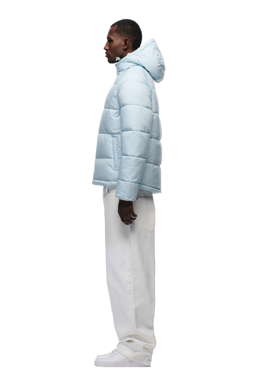 A man stands in profile wearing the 6PM PUFFER JACKET ICE BLUE with an adjustable hood, paired with white pants and sneakers, against a plain white background.