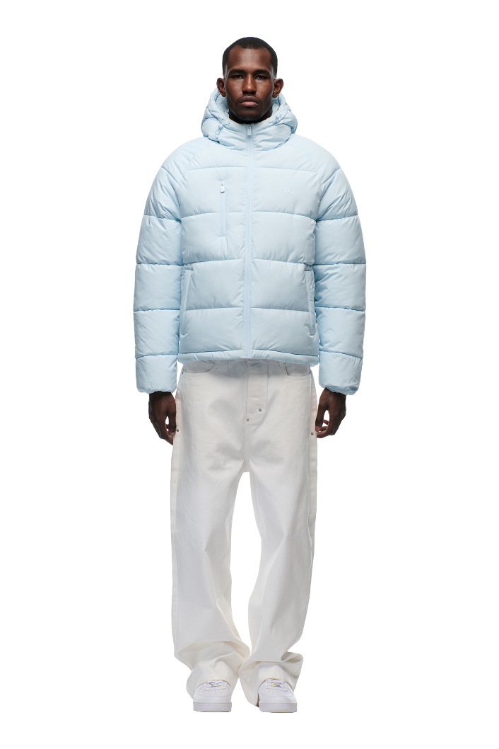 A man stands facing forward in a 6PM PUFFER JACKET ICE BLUE with an adjustable hood, paired with white baggy pants and white sneakers, against a plain white background.