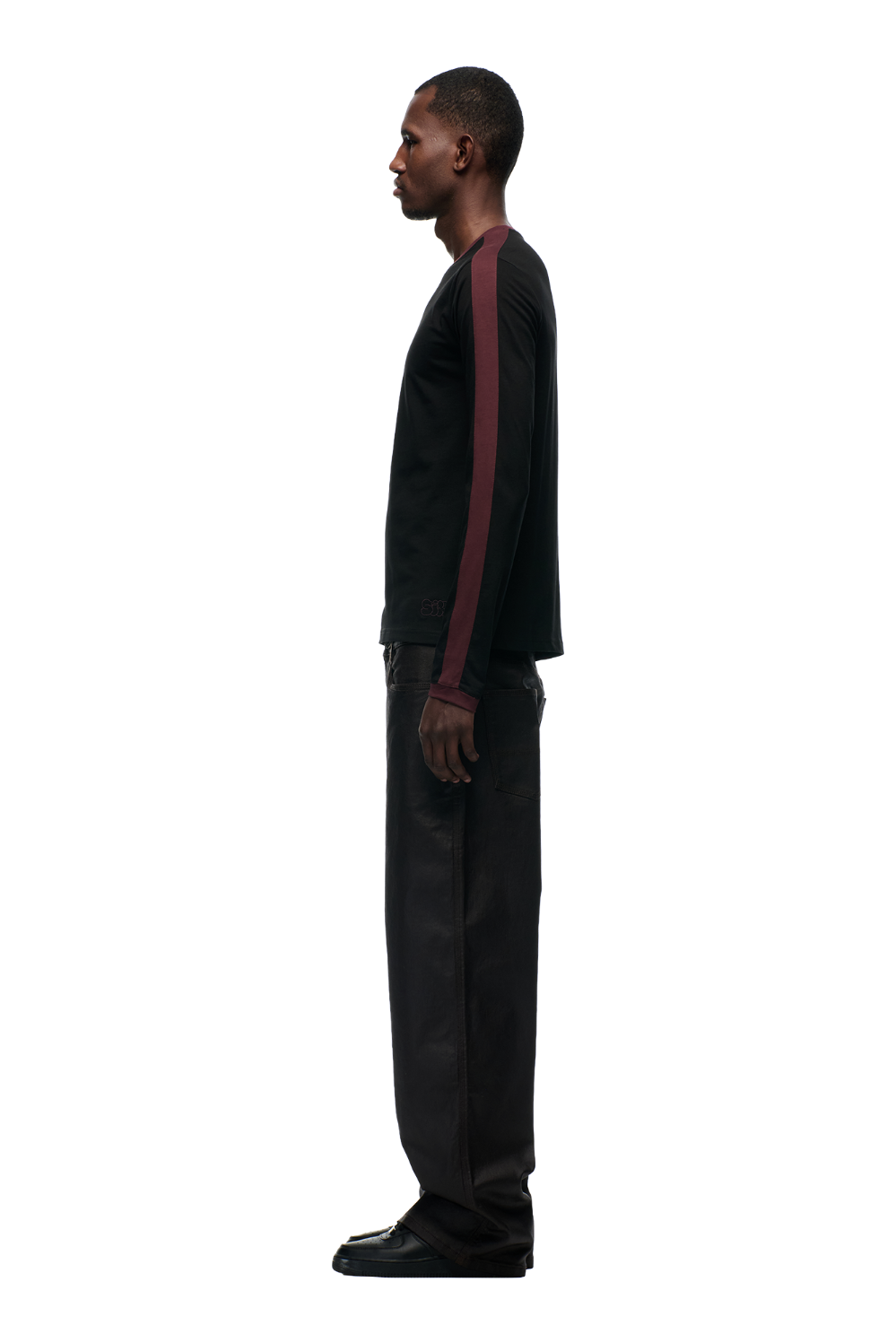 A man stands in profile facing left, wearing the 6PM GRAFFITI RINGER LS BLACK top, paired with loose black pants and black shoes, set against a plain light background.