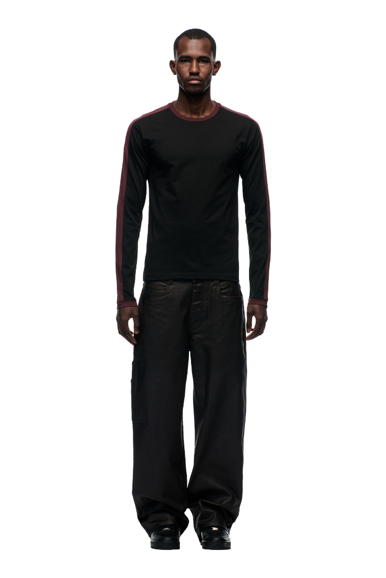 A man faces forward wearing the 6PM GRAFFITI RINGER LS BLACK, a black long-sleeve shirt with maroon stripes and color-block cuffs, paired with loose black pants against a white background.