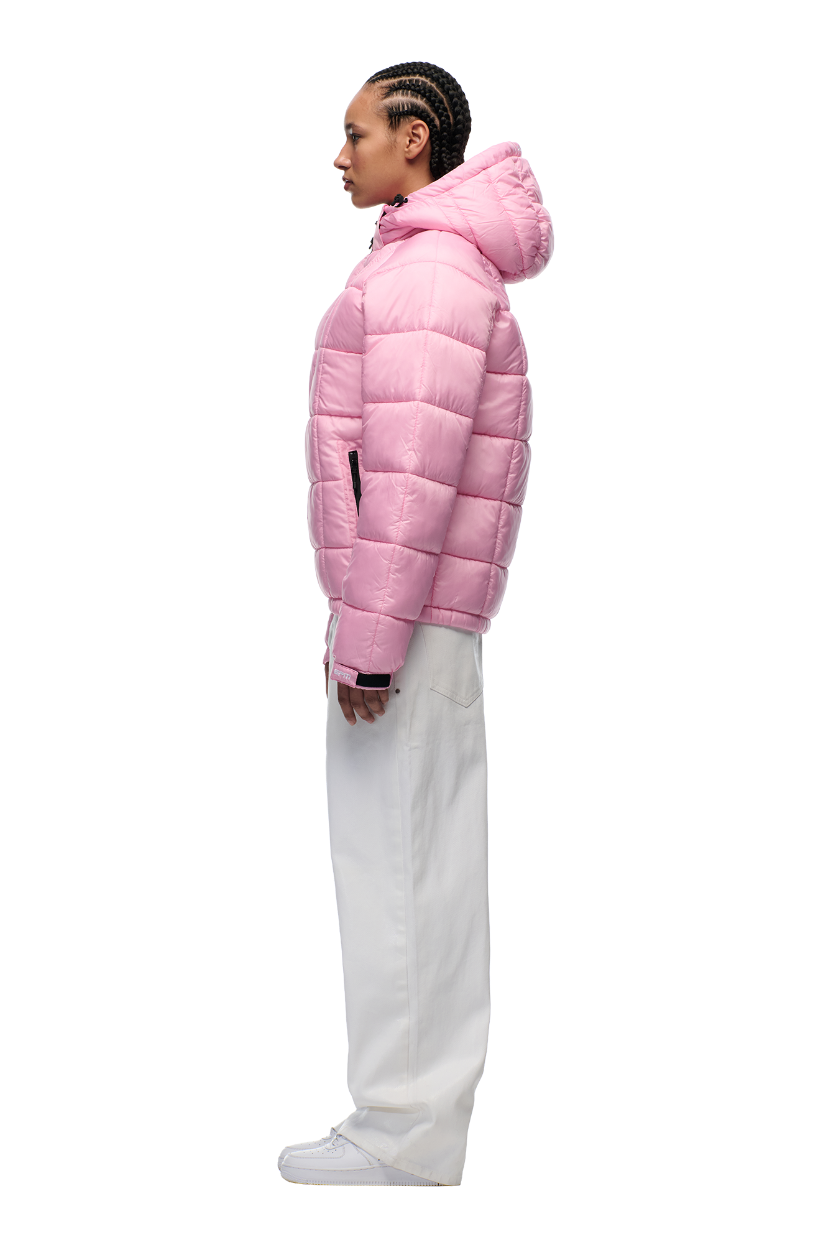 A person with braided hair stands in profile against a white background, wearing 6PM’s LIGHT PUFFER JACKET RECTANGLES PINK with an adjustable hood, paired with loose white pants and white sneakers.