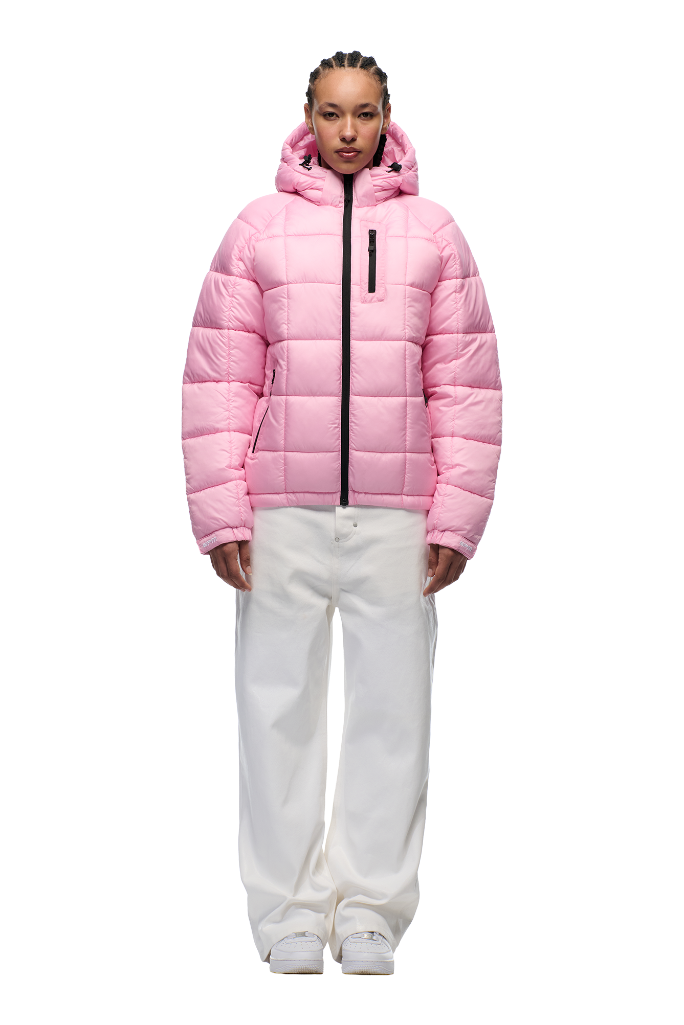 A person stands facing forward against a white background, wearing the 6PM LIGHT PUFFER JACKET RECTANGLES PINK with an adjustable hood and black zipper, paired with loose white pants and white shoes.