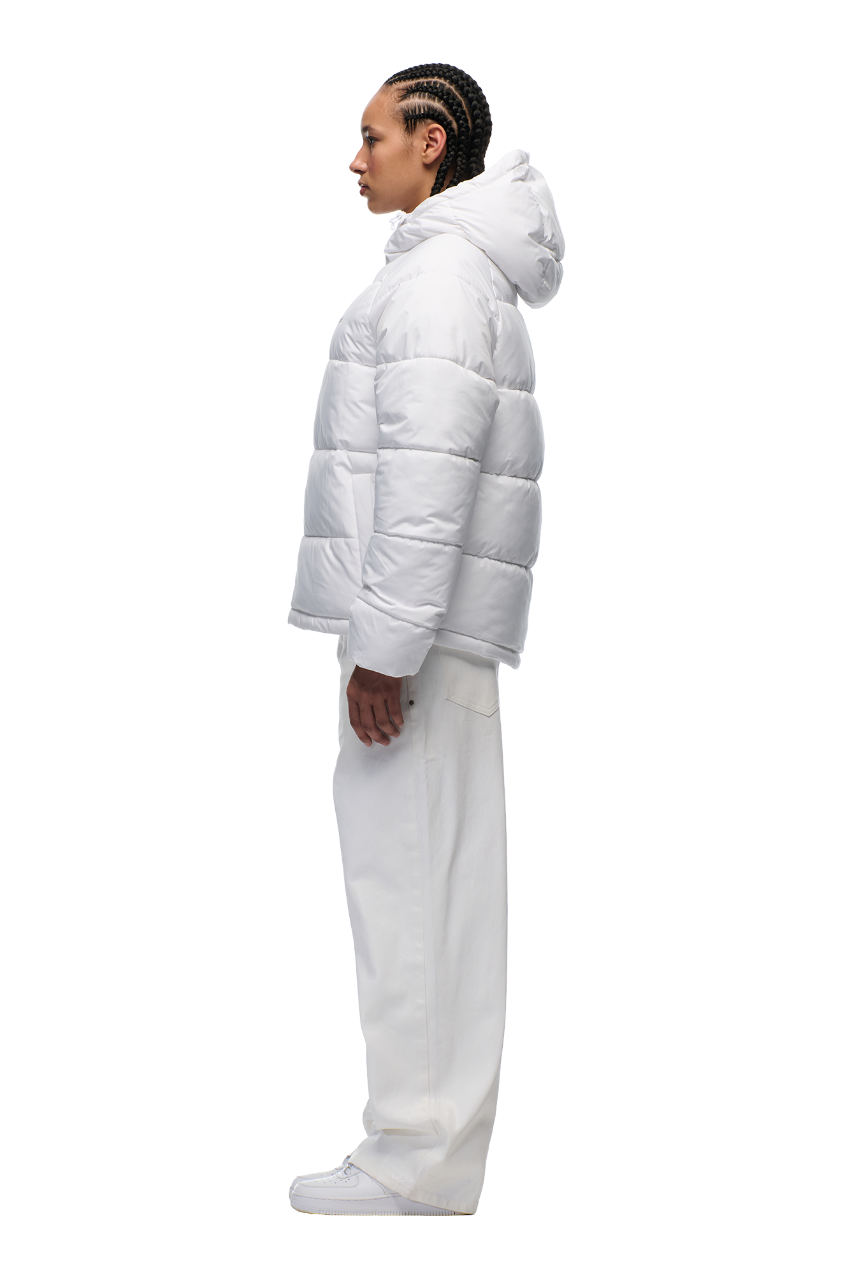 A person with braided hair stands in profile, dressed in all-white with loose pants and sneakers, showcasing the 6PM PUFFER JACKET WHITE—featuring water-repellent fabric and embroidered branding—against a plain white background.