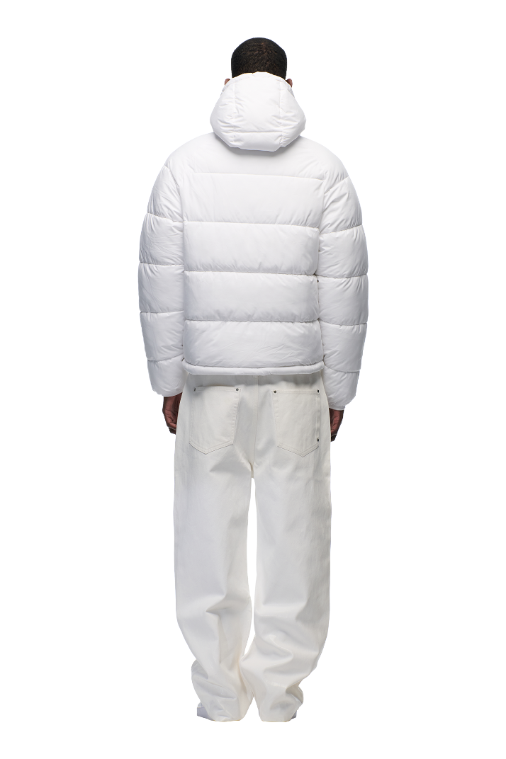 A person faces away, wearing the 6PM PUFFER JACKET WHITE featuring water-repellent fabric and an adjustable hood, paired with loose white pants, against a plain light background.