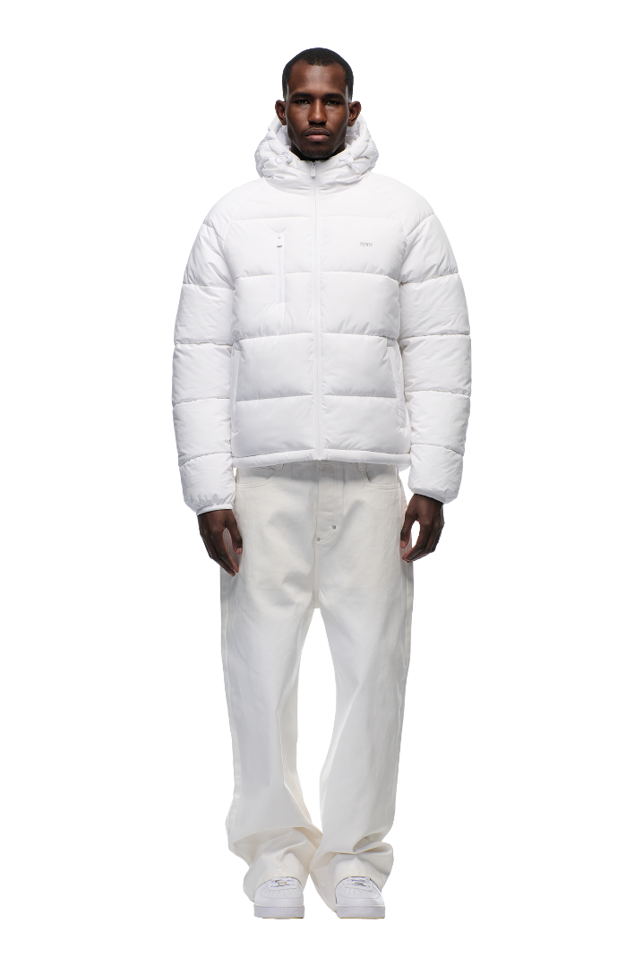 A man faces forward, wearing the 6PM PUFFER JACKET WHITE with an adjustable hood, paired with loose white pants and white sneakers, set against a plain light background.