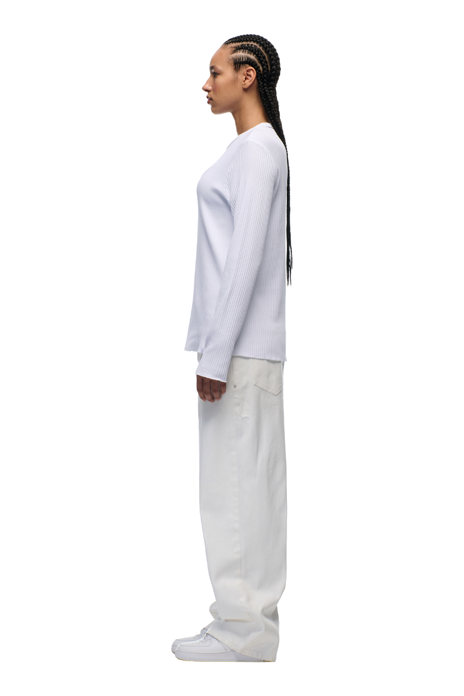 A person stands in profile against a white background, wearing the 6PM 6PM+ RIBBED LS WHITE top with long braids, loose white pants, and white shoes.