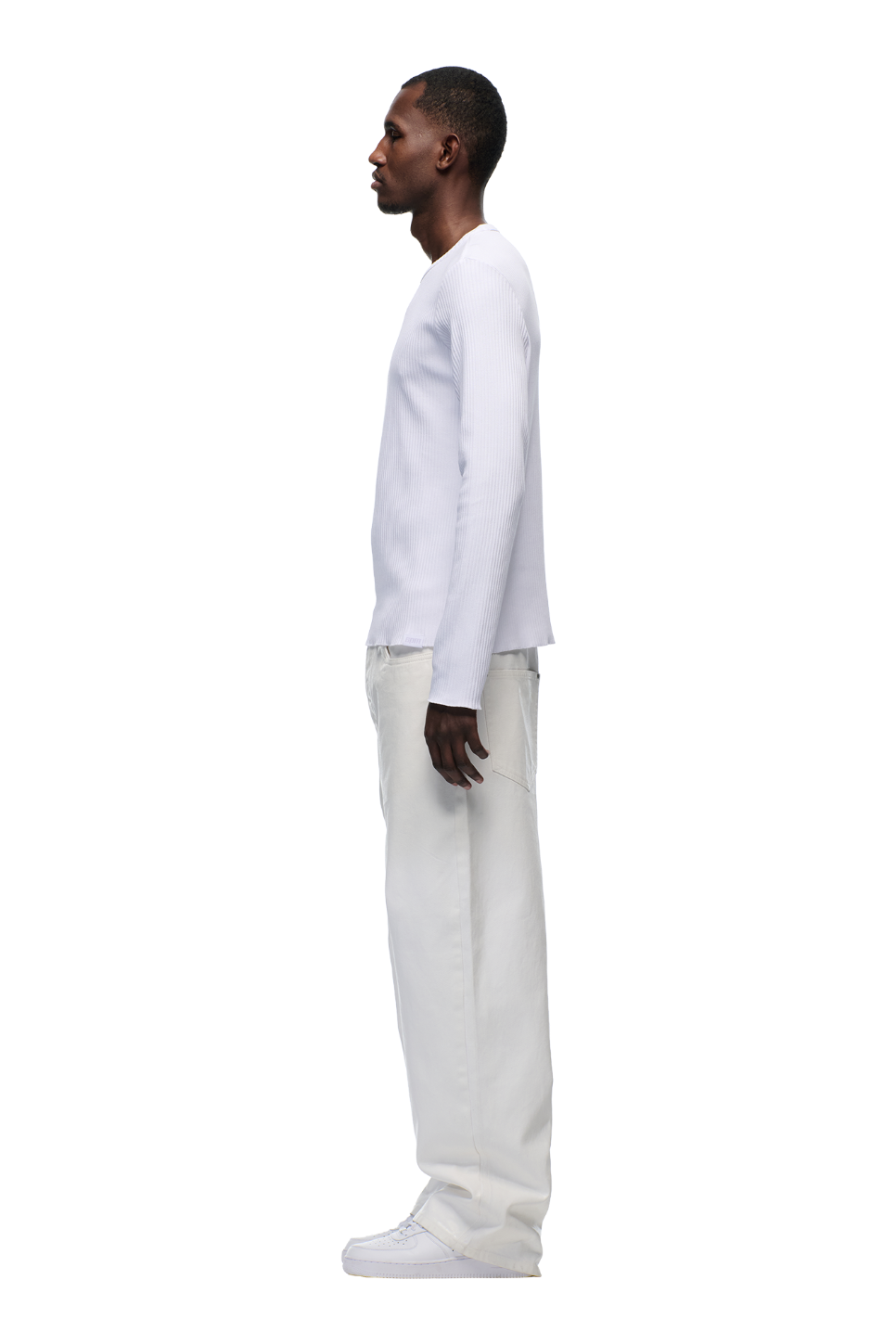 A man stands in profile facing left, dressed in the 6PM 6PM+ RIBBED LS WHITE top, loose white pants, and white sneakers with a tonal flag label, all set against a plain white background.