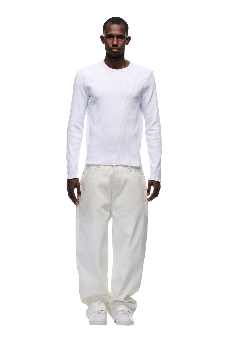 A man stands facing forward against a plain white background, wearing the 6PM 6PM+ RIBBED LS WHITE fitted long-sleeve top, loose white pants, and white sneakers.