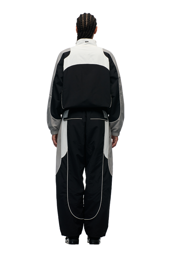 A person with braided hair is shown from the back, wearing the 6PM EVERYDAY TRACK JACKET BLACK, a black, white, and gray nylon jacket with mesh lining, against a plain white background.