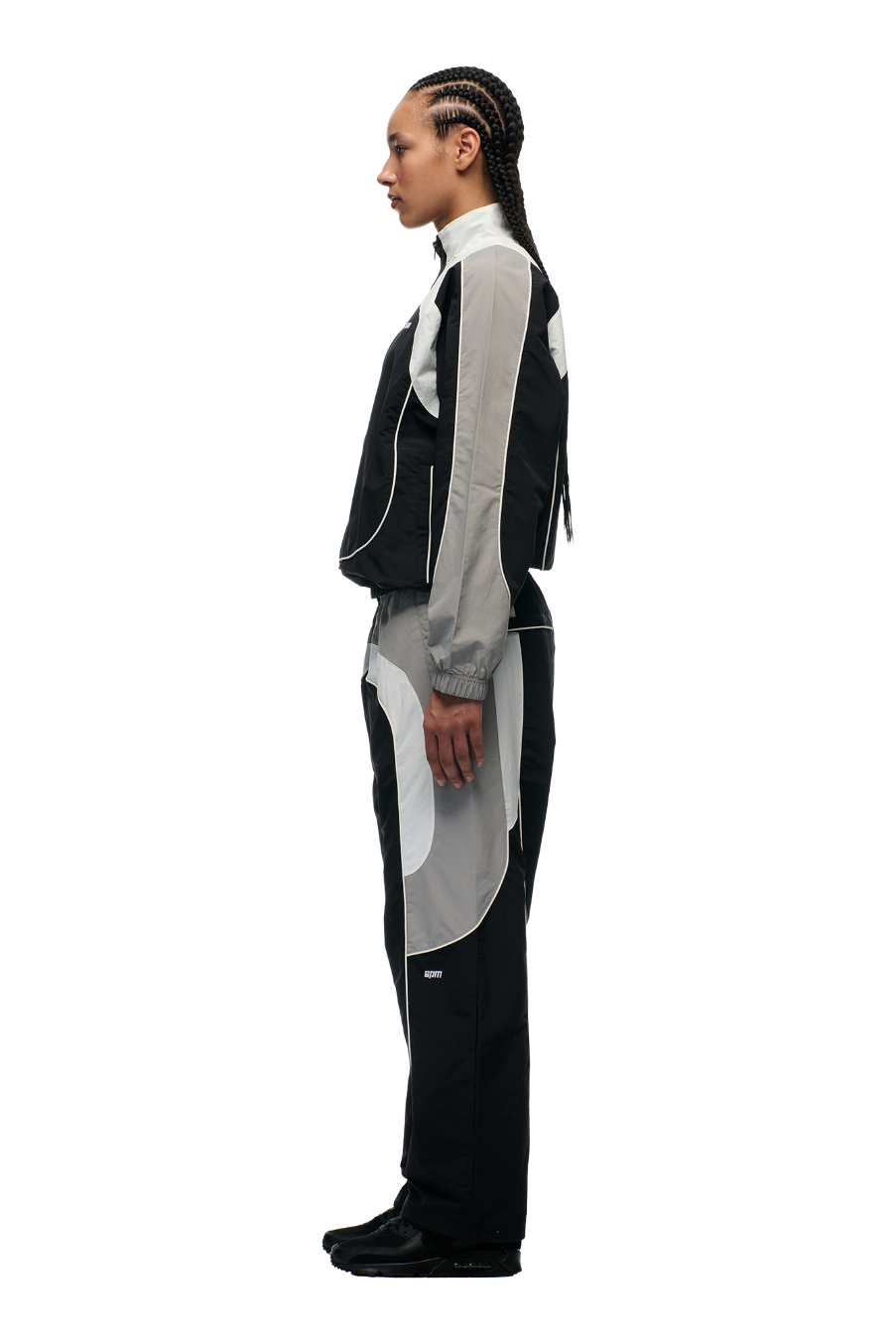 A person with braided hair stands in profile against a white background, wearing the 6PM EVERYDAY TRACK JACKET BLACK with matching pants, both featuring embroidered logos, paired with black shoes.