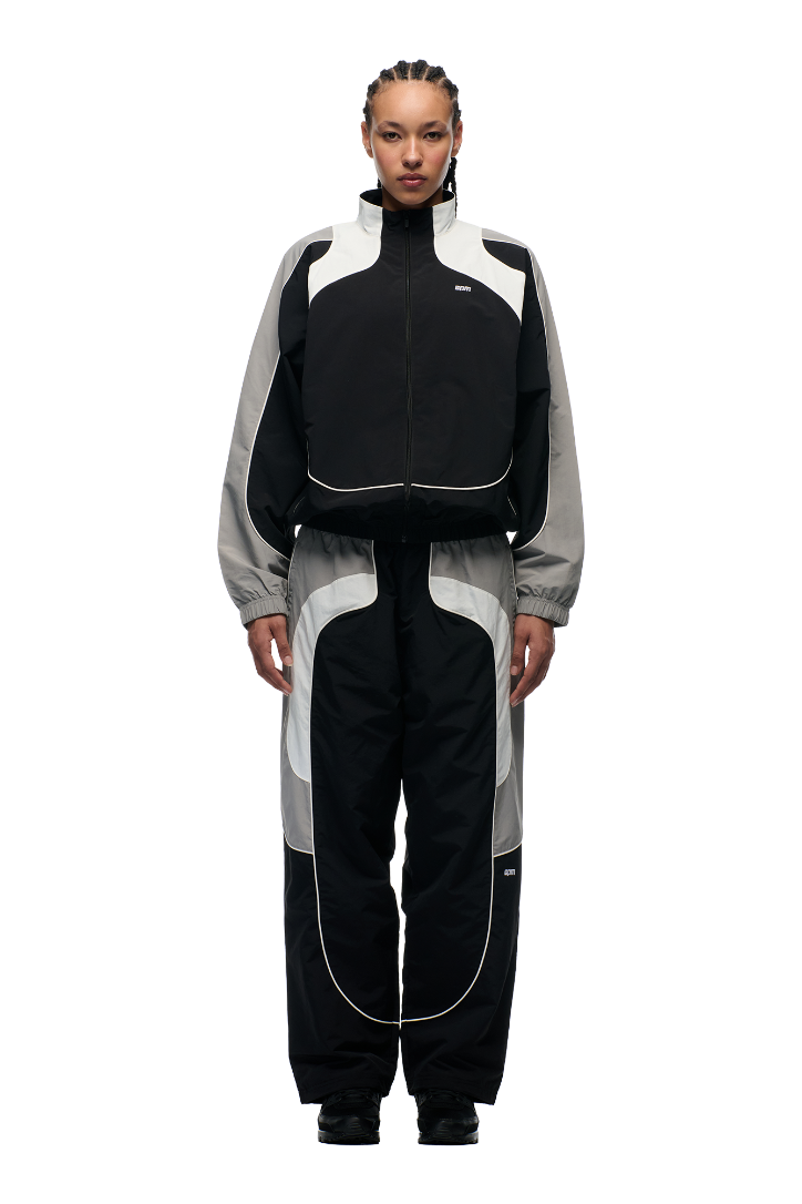 A person with braided hair faces forward, modeling the 6PM EVERYDAY TRACK JACKET BLACK with matching pants and black shoes, standing against a plain white background.