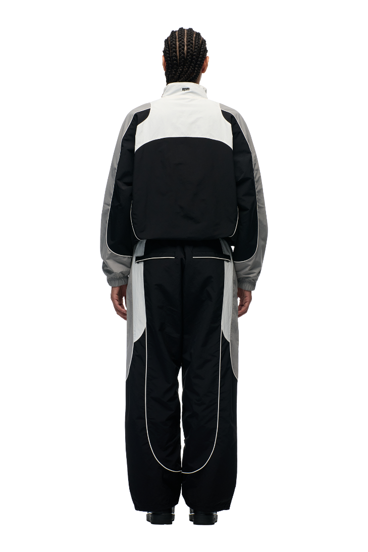 A person with braided hair faces away, wearing the 6PM EVERYDAY TRACK JACKET BLACK—loose-fitting with mesh lining, white piping, and subtle embroidered logos—paired with matching pants, set against a plain light background.