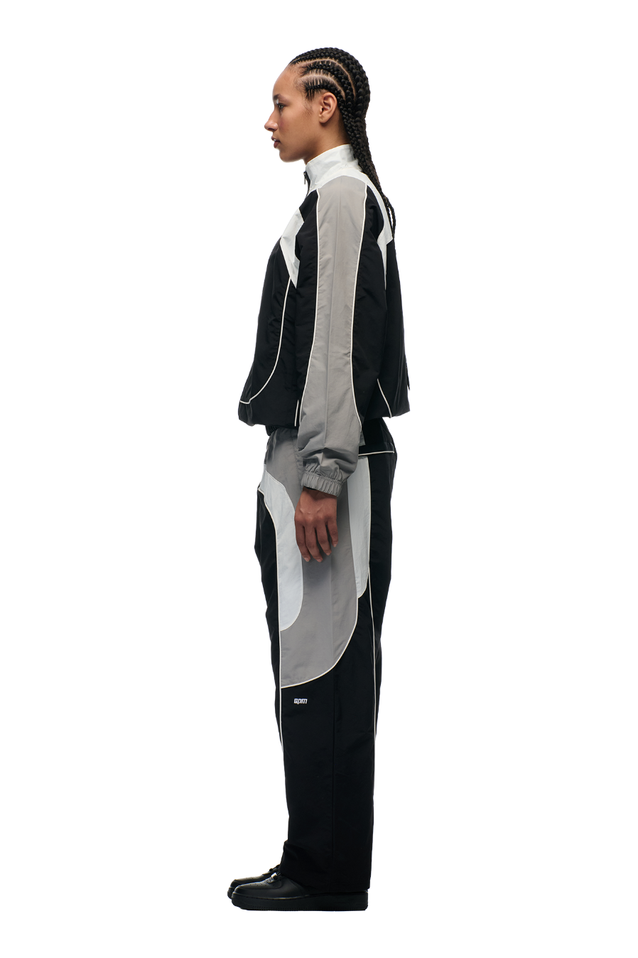 A person with braided hair, in profile, wears the 6PM EVERYDAY TRACK JACKET BLACK with embroidered logos and black shoes, standing against a plain white background.